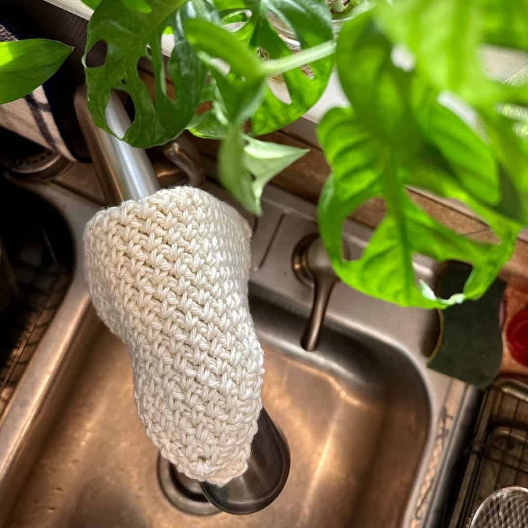 Hand-knit moss stitch crochet washcloth hanging on kitchen faucet with green plants.