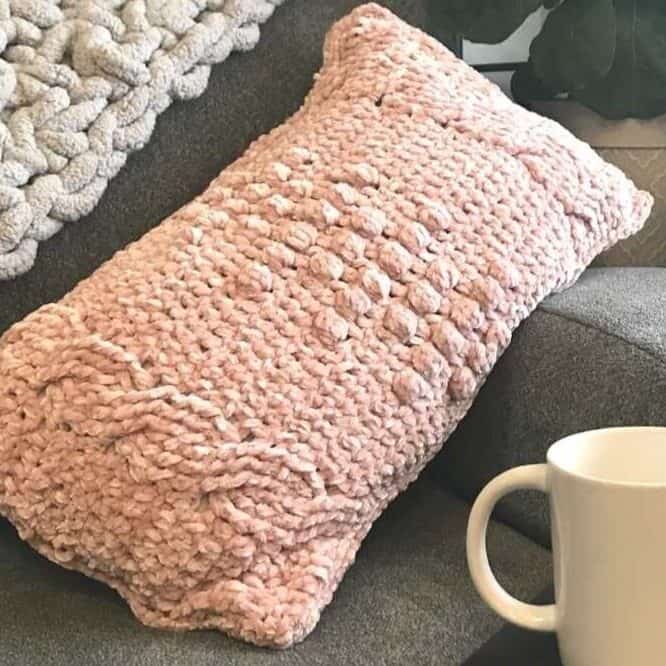 Pink chunky knit decorative pillow on a gray sofa with a white mug nearby.
