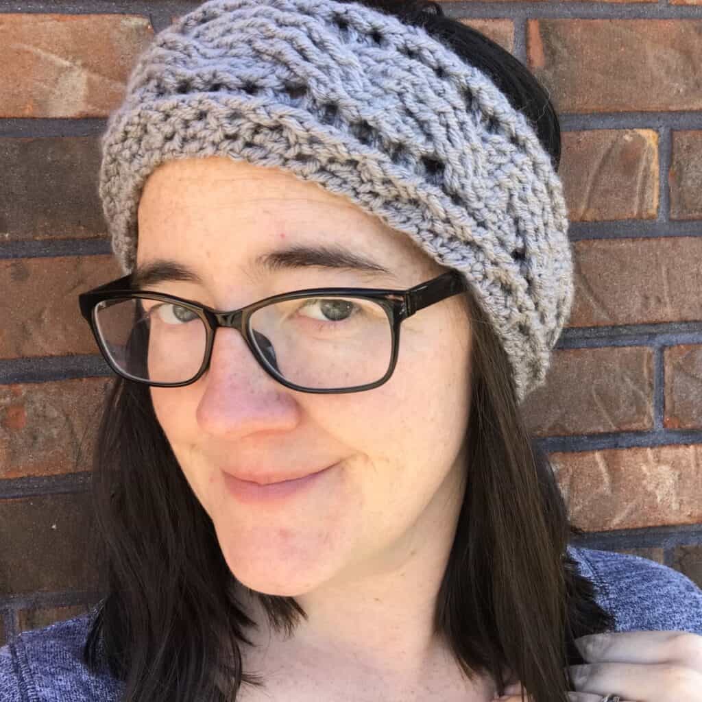 Woman with glasses wearing a knitted headband, smiling outdoors against a brick wall background.