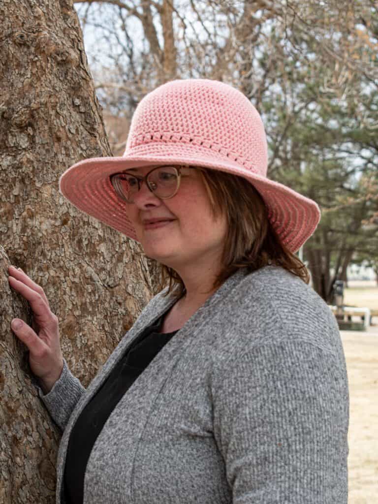 Outdoor portrait of a woman wearing a pink sun hat and glasses, standing beside a tree.
