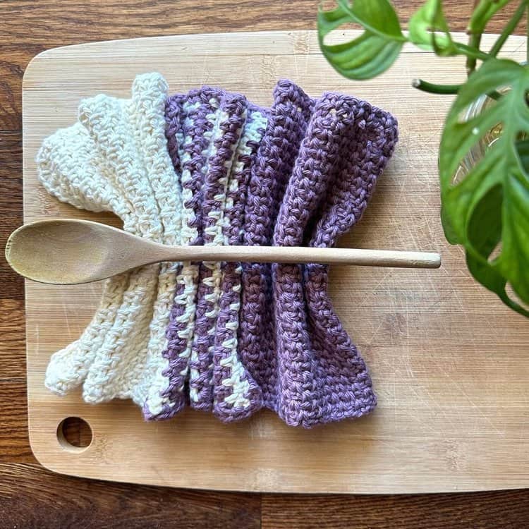 Crochet dishcloths in white and purple with a wooden spoon on a wooden cutting board.