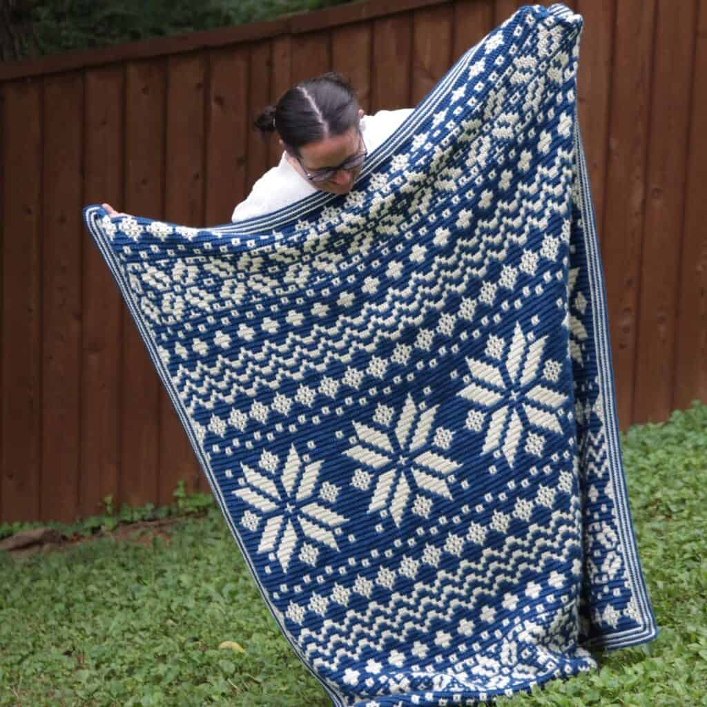 Woman holding up a blue and white mosaic crochet afghan. The blanket is winter/Christmas-themed.