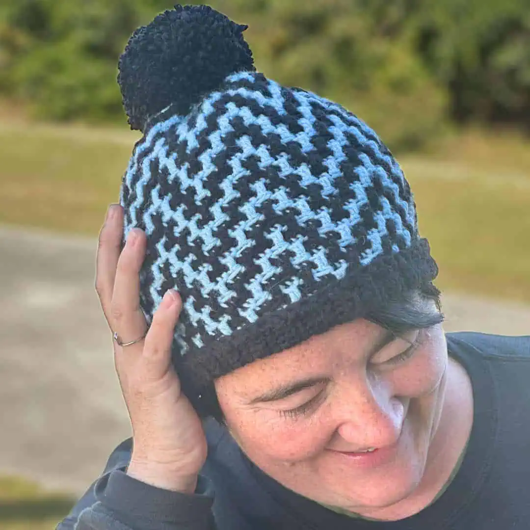 Close-up of a woman wearing a blue and black mosaic zigzag crochet hat with pom-pom.