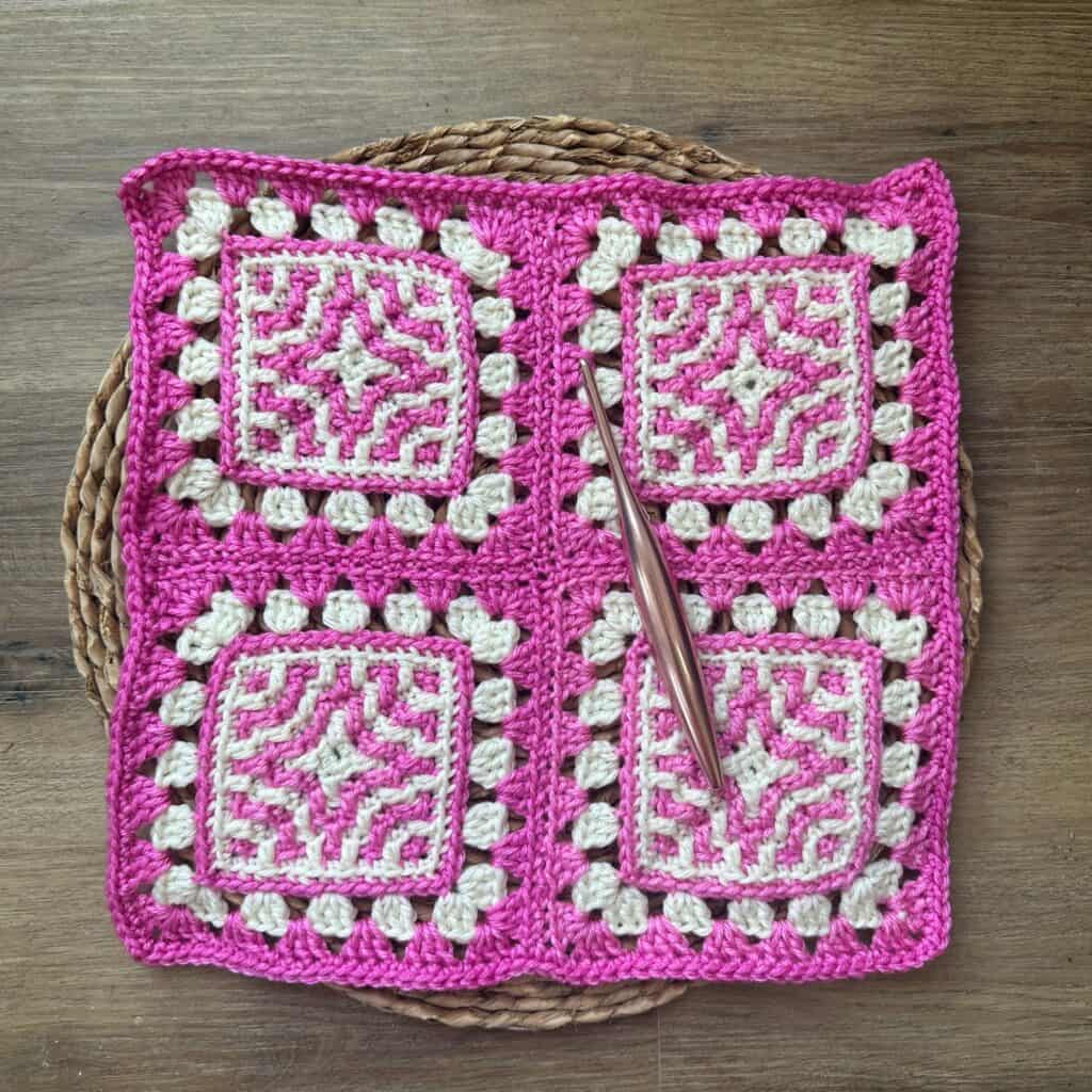 Pink and white crocheted granny square blanket with a crochet hook on a wooden surface.