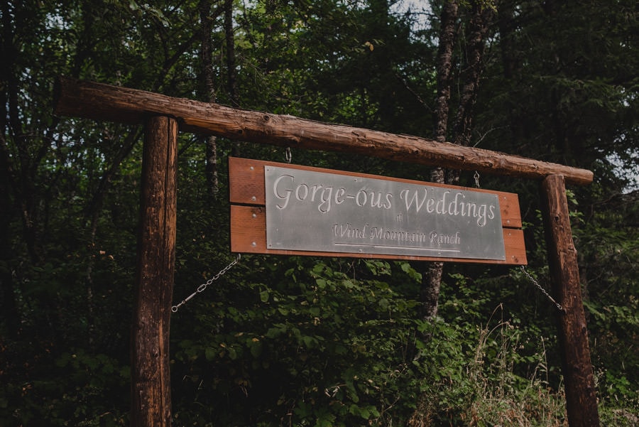 A wooden sign with Gorge-ous Weddings written on it is positioned at Wind Mountain Ranch, surrounded by lush green trees.