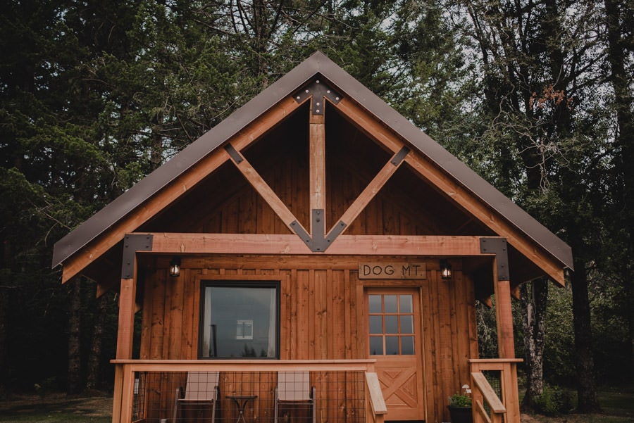An A-frame cabin with rustic wooden textures is nestled among tall trees on a peaceful mountain ranch. The cabin has a cozy porch illuminated with soft lights.