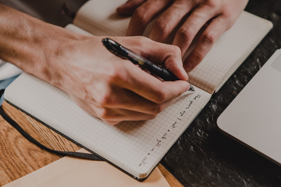 Hands writing notes in a grid notebook with a pen next to a laptop on a dark surface, with an open window in the background showing mountains.