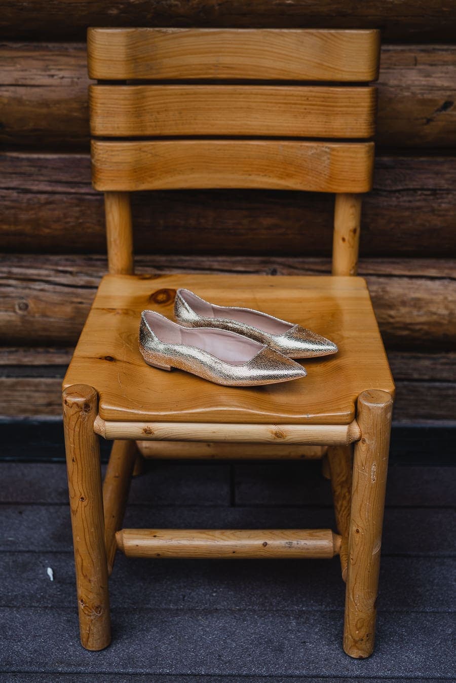A pair of gold flat shoes is placed on a wooden chair, with a background of log cabin walls.