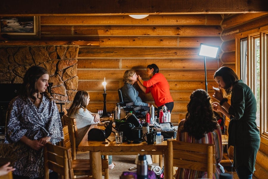 In a cozy rustic room with wooden walls, preparations are underway for a wedding. A person is seated while having their makeup applied, surrounded by photography equipment capturing the scene.
