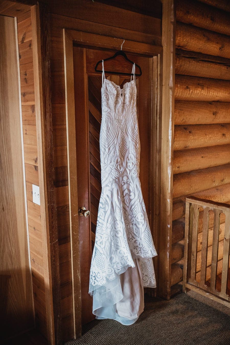 A white lace wedding dress hangs elegantly on a wooden door in a rustic cabin, showcasing the charm of the setting.