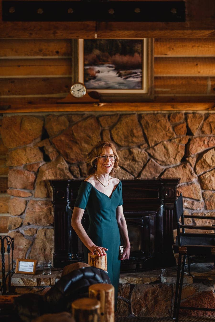 A woman in a green dress smiles while standing in a rustic room with a stone wall and fireplace.