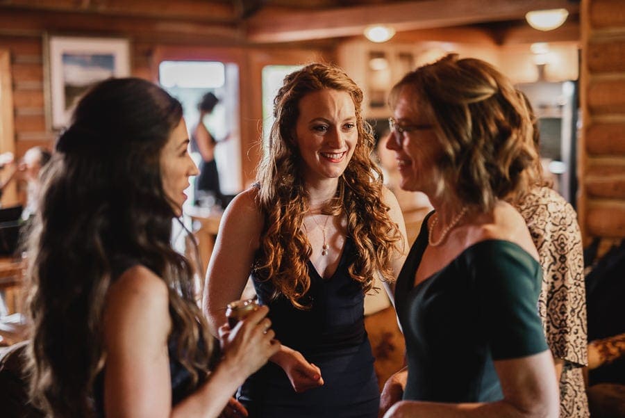 Three women in conversation at a warmly lit gathering, with wooden walls creating a rustic backdrop.