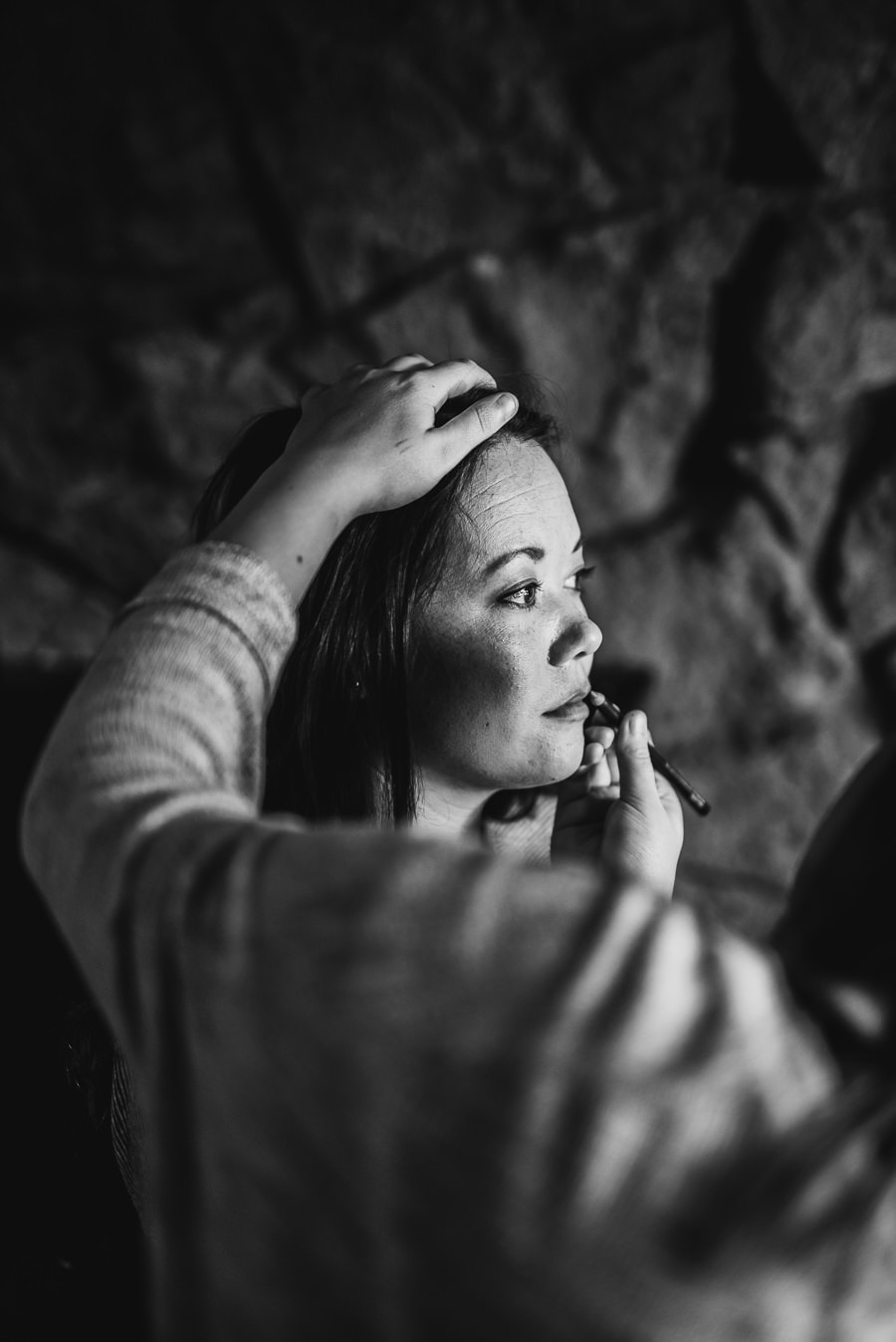 A person receives flawless makeup against a textured, stone-like backdrop. Hands gently cradle their head and chin.