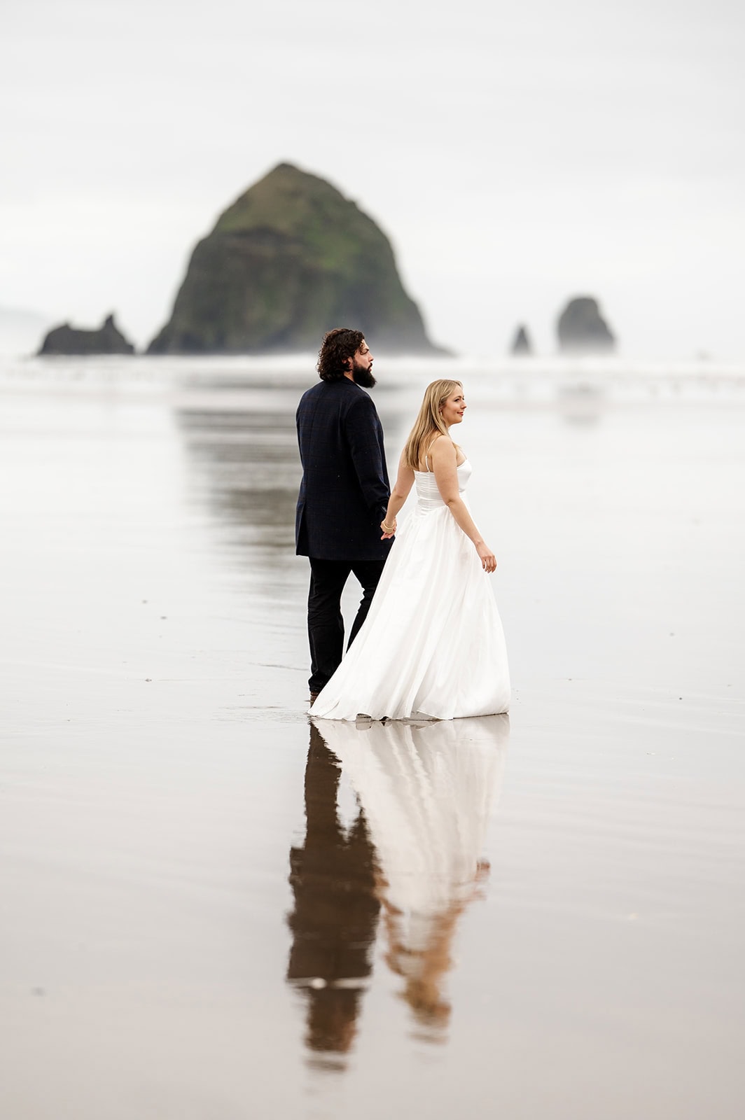 A couple holds hands while walking along the wet sand of a beach with large rock formations in the background, a wonderful reason to elope.