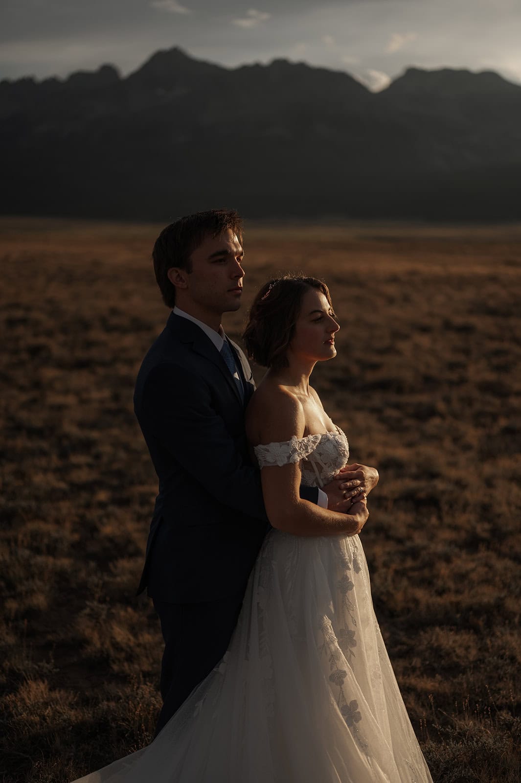 A bride in a white off-the-shoulder wedding gown and a groom in a dark suit standing together in a field at sunset, with the Sawtooth Mountains in the background of their elopement.