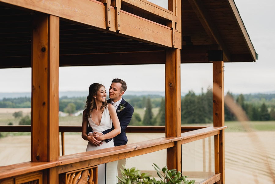 A couple smiles and embraces on a wooden balcony at Scholls Valley Lodge, with wedding photos displayed around them. The scenic landscape of rolling hills and trees is visible in the background.
