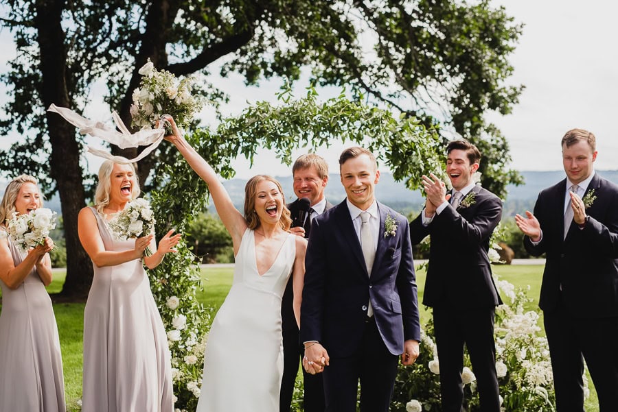 A joyful wedding ceremony at Scholls Valley Lodge shows a happy couple celebrating with bridesmaids and groomsmen under a floral arch. The image captures their smiles and the vibrant flowers adorning the arch, set against a sunny outdoor backdrop.