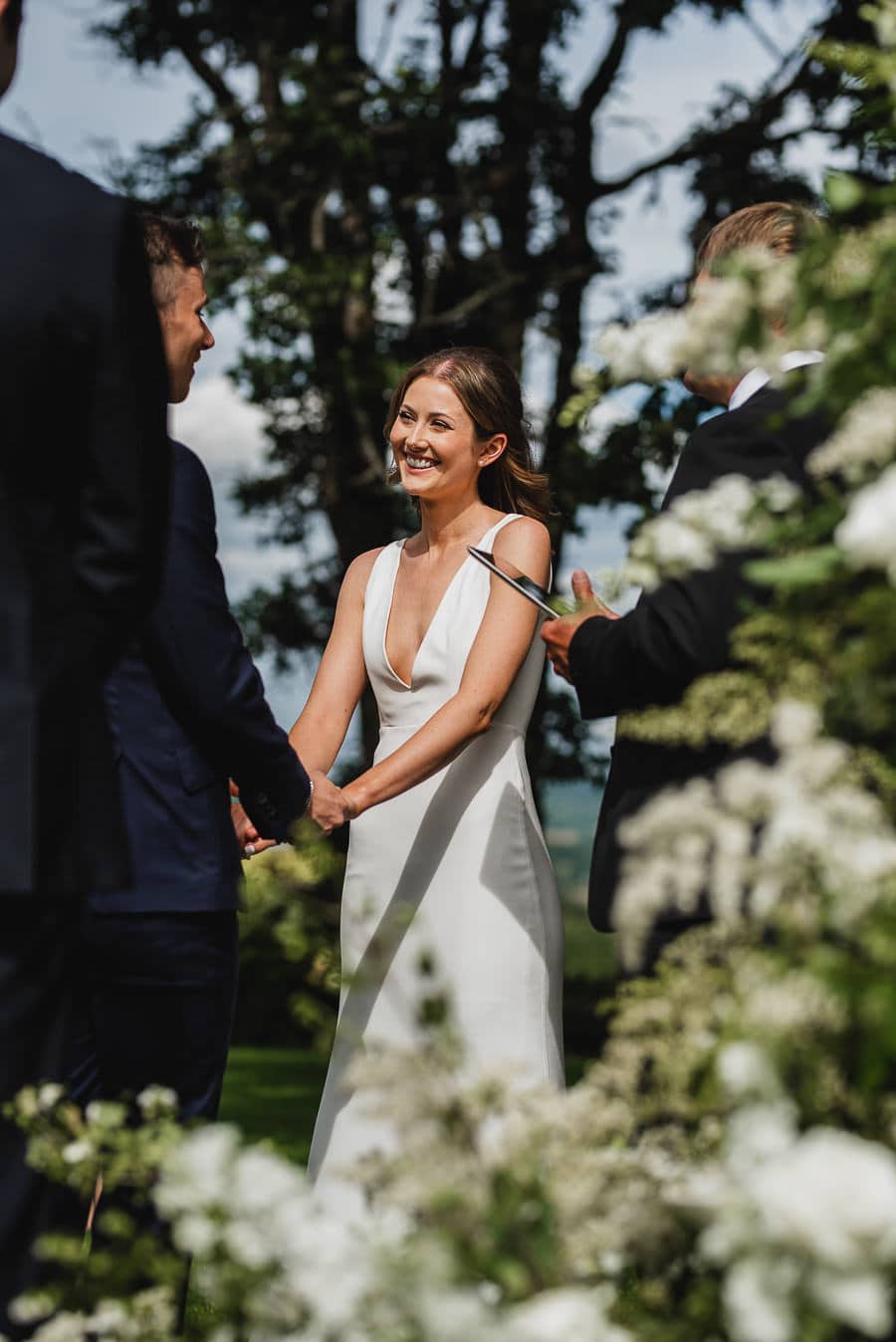 Bride and groom holding hands and smiling outdoors at Scholls Valley Lodge, with greenery and white flowers in the background.