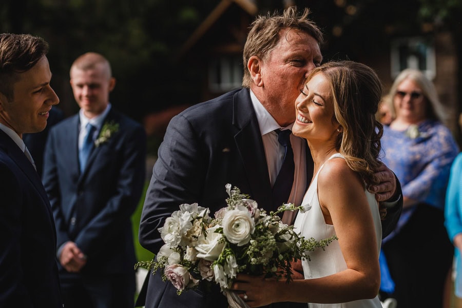 A woman in a white dress holding flowers smiles as a man kisses her cheek during an outdoor ceremony at Scholls Valley Lodge.