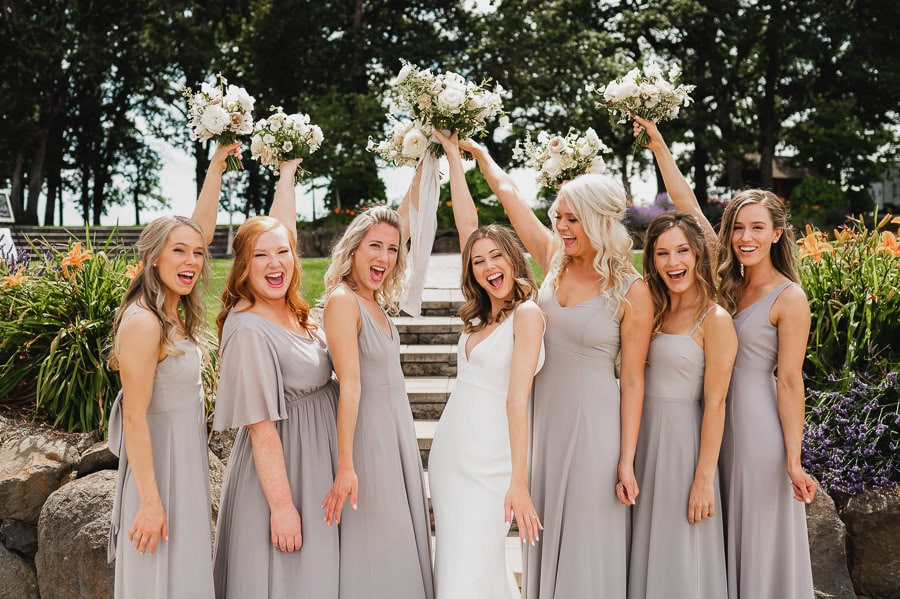 Bridesmaids in gray dresses and a bride in white joyfully raise their bouquets while posing outdoors at Scholls Valley Lodge, creating a vibrant wedding photo.