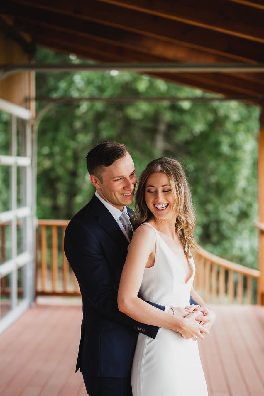 A smiling couple embraces on the wooden porch of Scholls Valley Lodge, surrounded by lush greenery, in a perfect wedding photo.