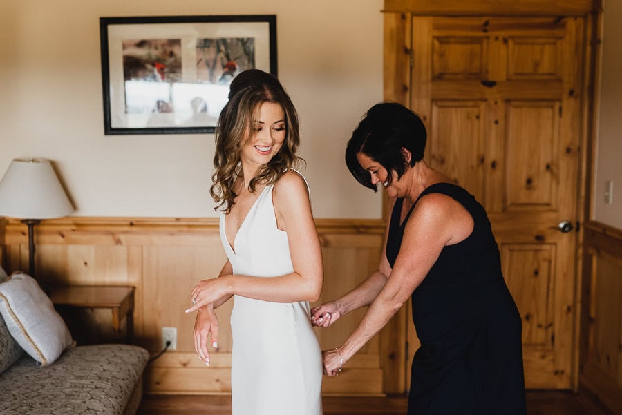 In a cozy wooden room at Scholls Valley Lodge, a woman adjusts the wedding dress of another, both smiling warmly as they get ready for their special day.
