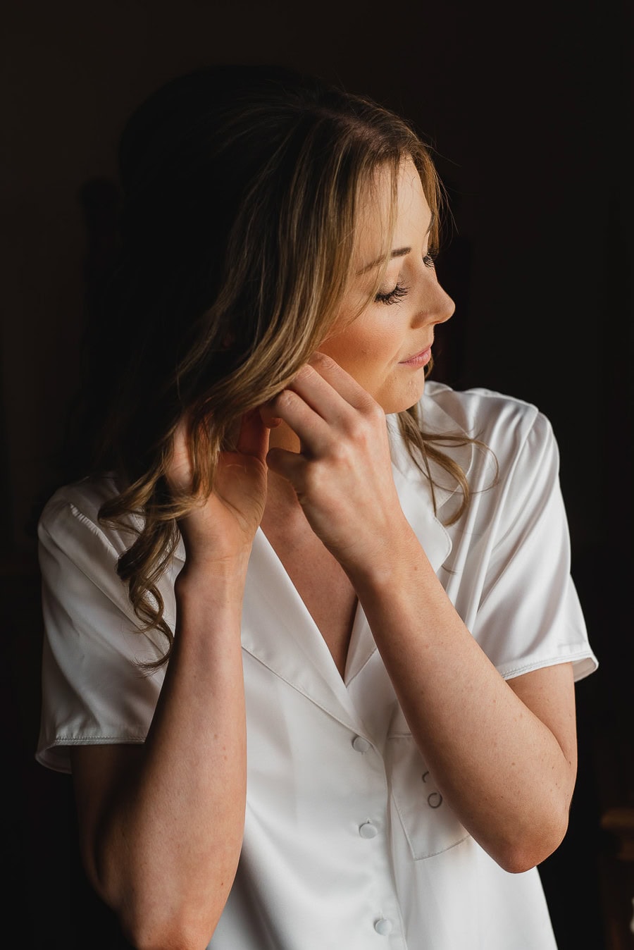 A woman in a white shirt adjusts her earring and gazes thoughtfully to the side against a dark background, surrounded by wedding photos.
