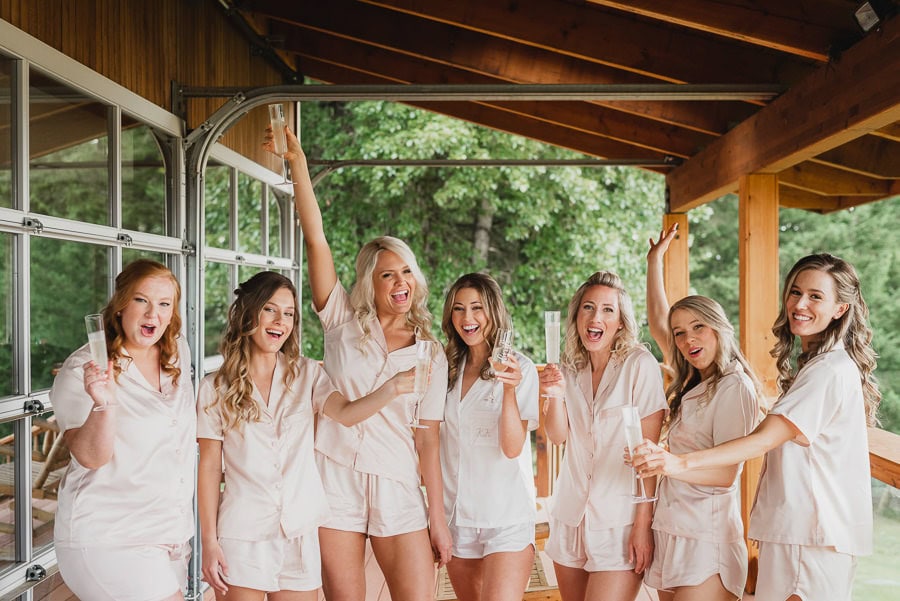 Seven women wearing matching pajamas enjoy drinks together on a porch at Scholls Valley Lodge, with lush greenery in the background.