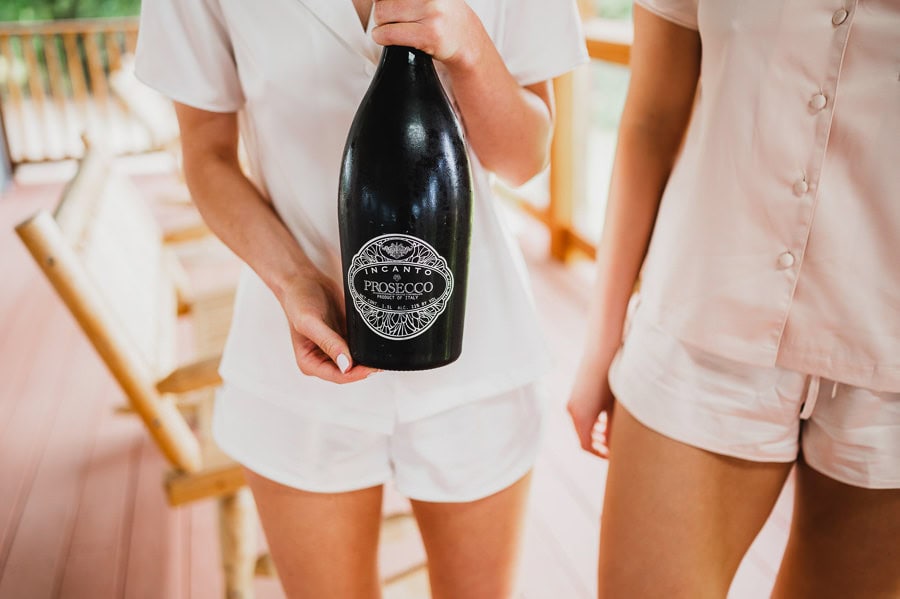 Two people in matching pajamas standing on a wooden deck, holding a large bottle of Prosecco and smiling.