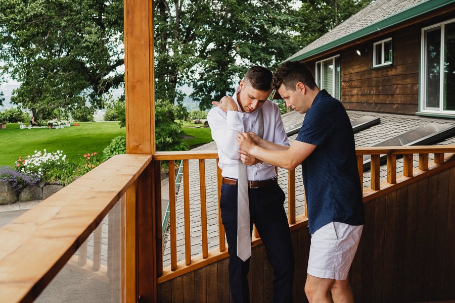 Two men stand on a wooden porch at Scholls Valley Lodge, with one adjusting the others tie. The background features a green lawn and trees.