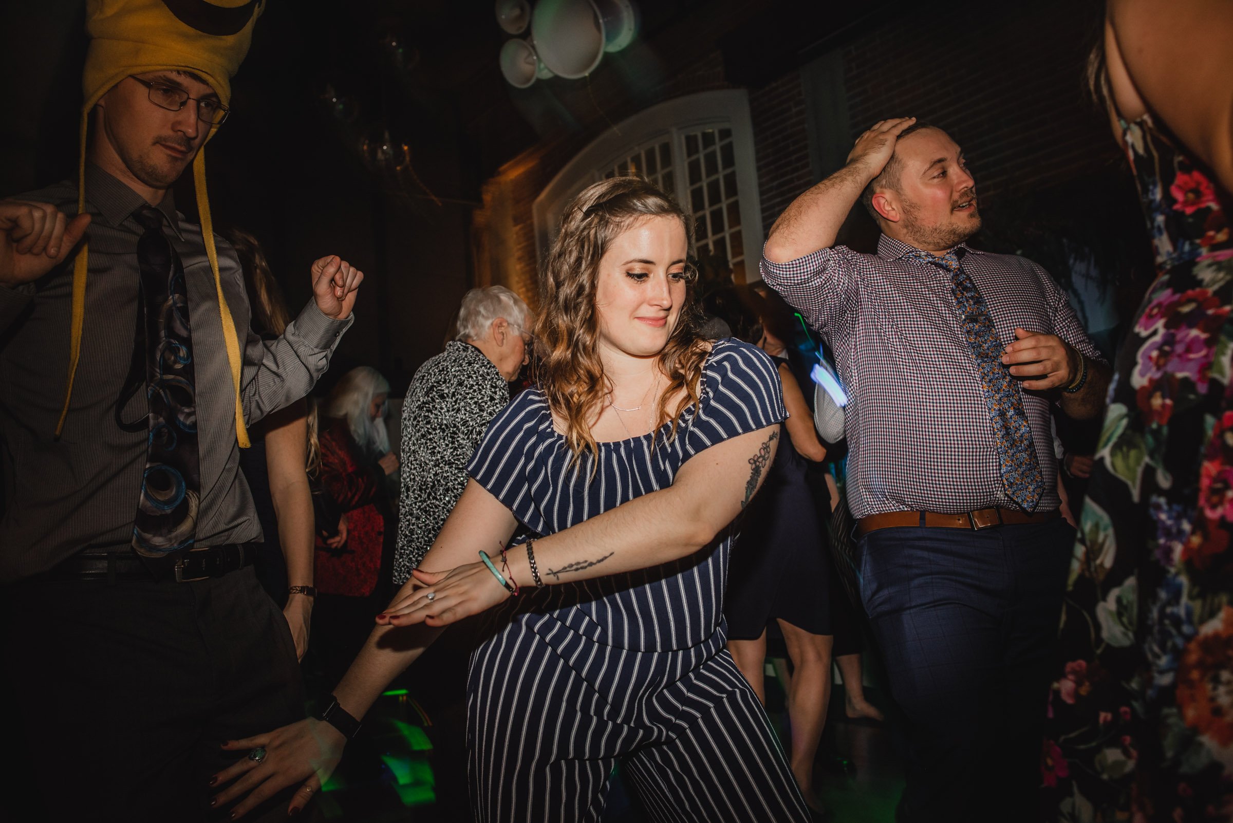 People dancing energetically in a dimly lit room, dressed in a mix of casual and formal attire as if captured in lively wedding photos at The Evergreen.