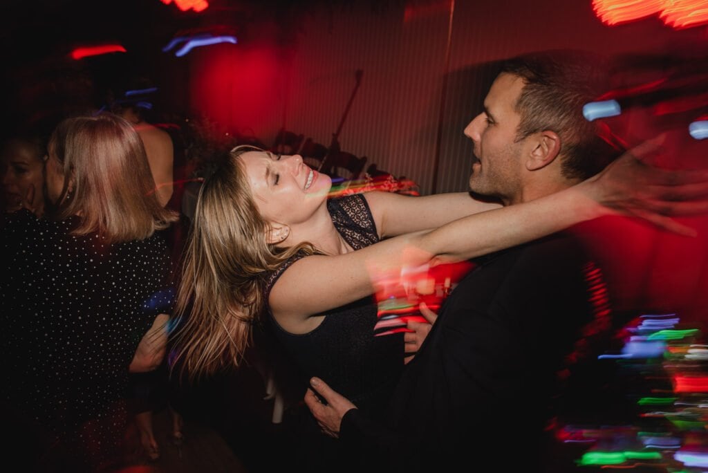 A joyful couple dances at a lively party with colorful lights and a blurred motion effect, as if captured in vibrant wedding photos at The Evergreen.
