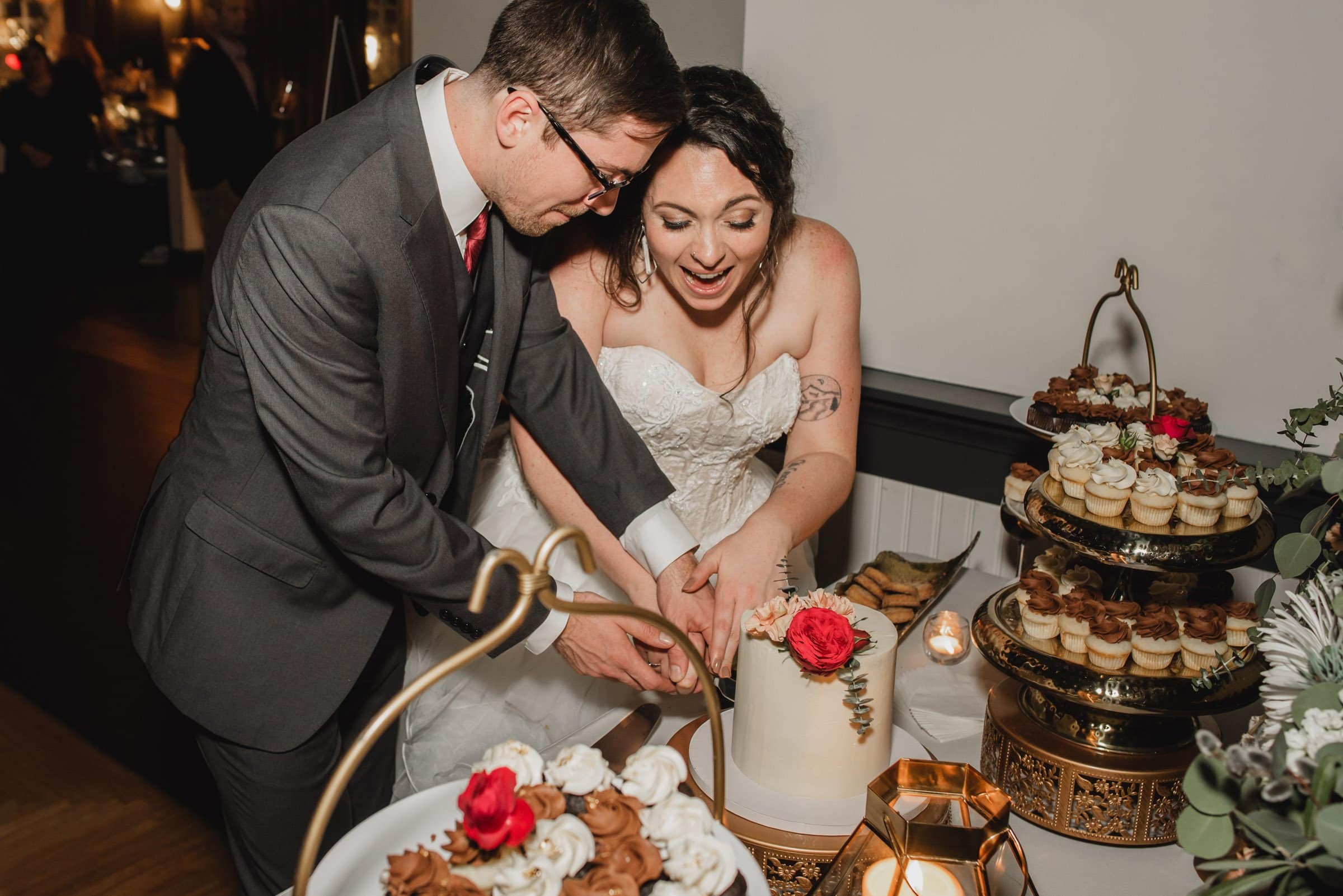 Bride and groom smiling and cutting a decorated cake at their wedding reception, surrounded by cupcakes. The joyous moment is captured beautifully in their wedding photos at The Evergreen.