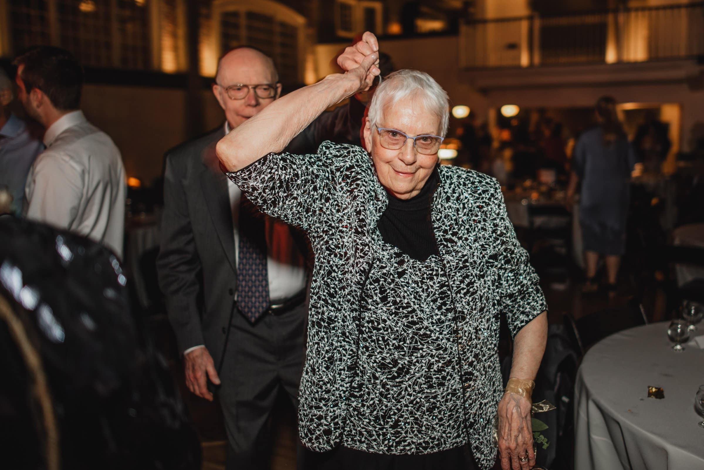 An older woman smiles and dances at an indoor event, joyfully holding hands with a man in the background. It seems like a celebration captured in wedding photos at The Evergreen.