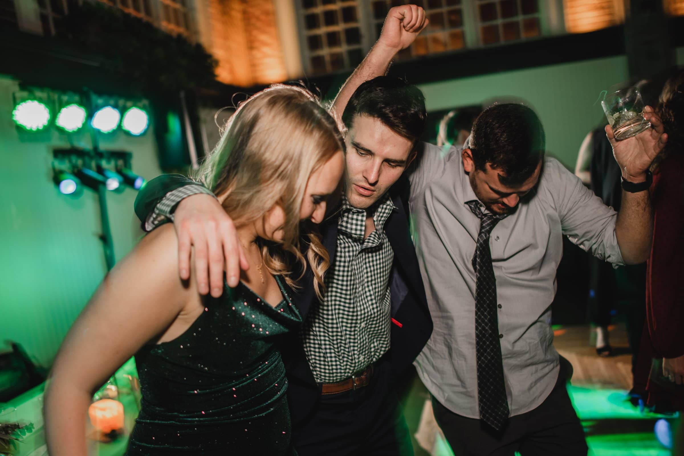 Three people enjoying a dance floor, arms around each other, with colorful lights in the background, capturing the joy of wedding photos at The Evergreen.
