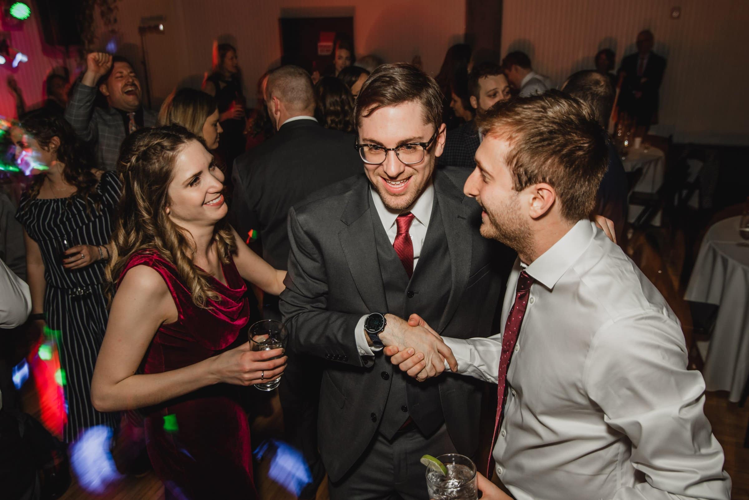 People smiling, shaking hands, and holding drinks at a lively indoor event, capturing the joy often seen in wedding photos at The Evergreen.