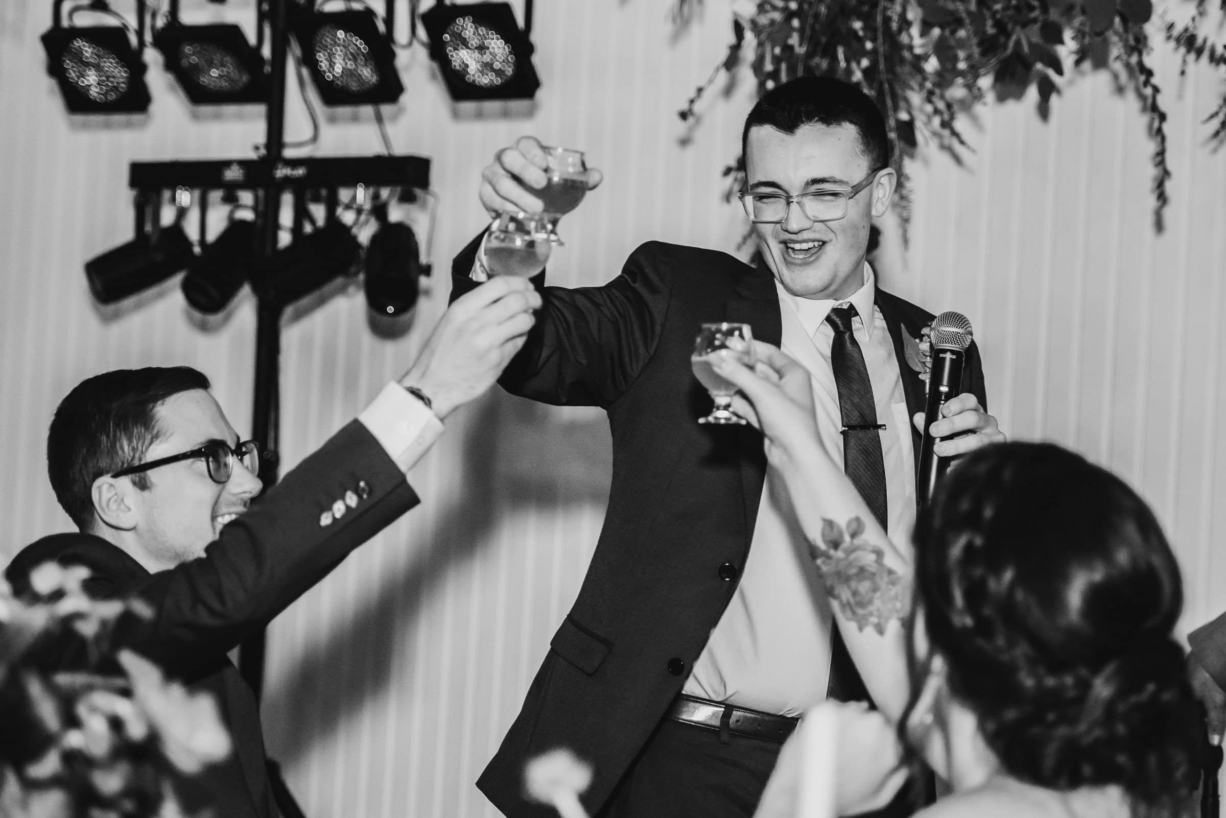 A man gives a toast with a microphone as others raise their glasses at a formal event. Captured in black and white, this wedding photo captures the elegance of the celebration at The Evergreen.