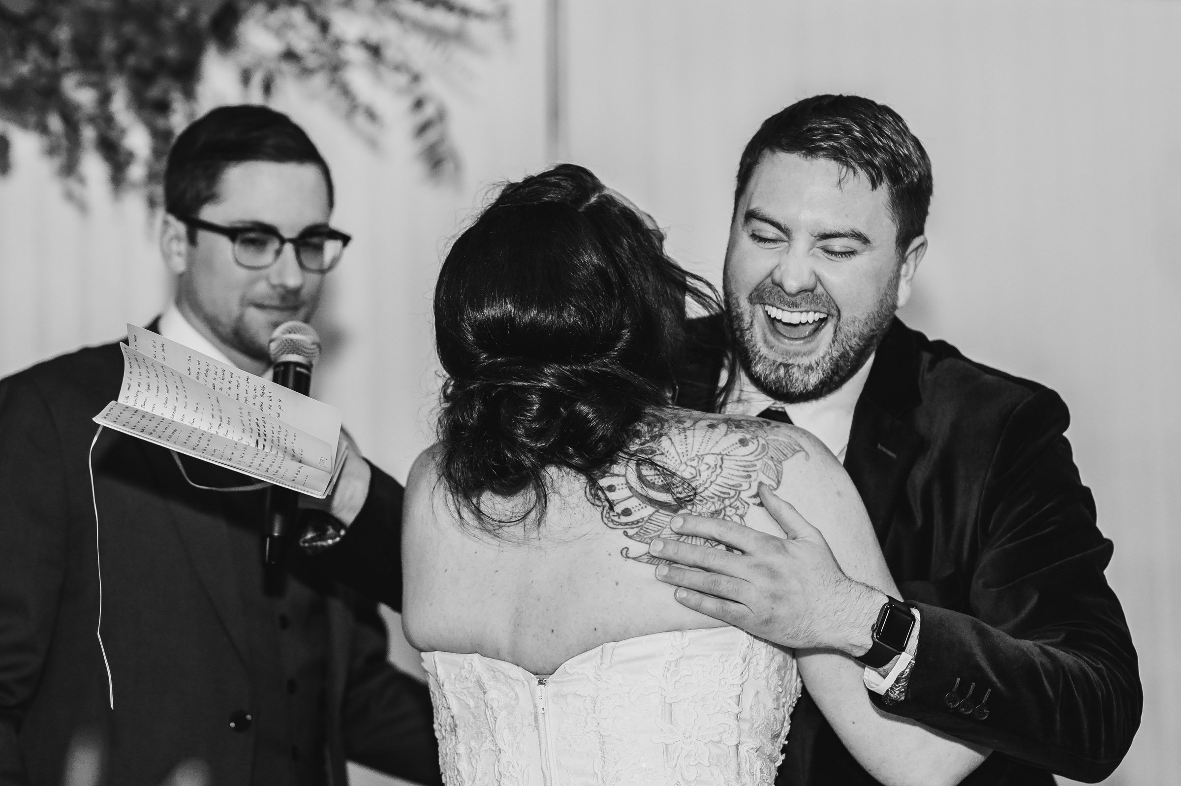 The bride and groom joyously embrace during their wedding at The Evergreen, while a man holding a microphone stands close by, capturing the moment for their wedding photos.