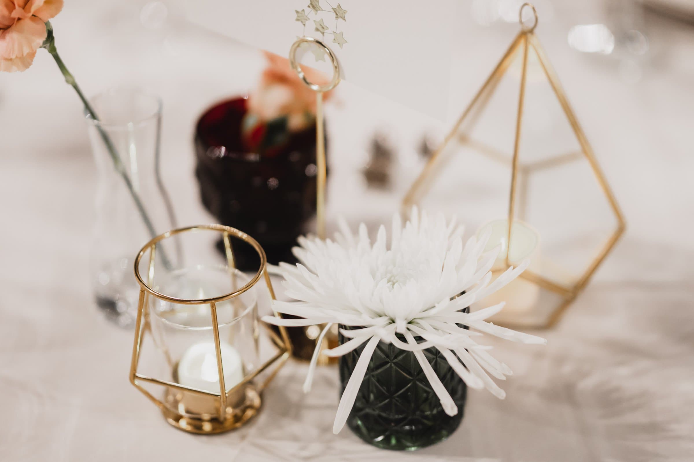 This elegant table arrangement features a white floral centerpiece complemented by gold geometric candle holders and a dark red glass vase. Perfect for capturing stunning wedding photos at The Evergreen, it adds a touch of sophisticated charm to any setting.