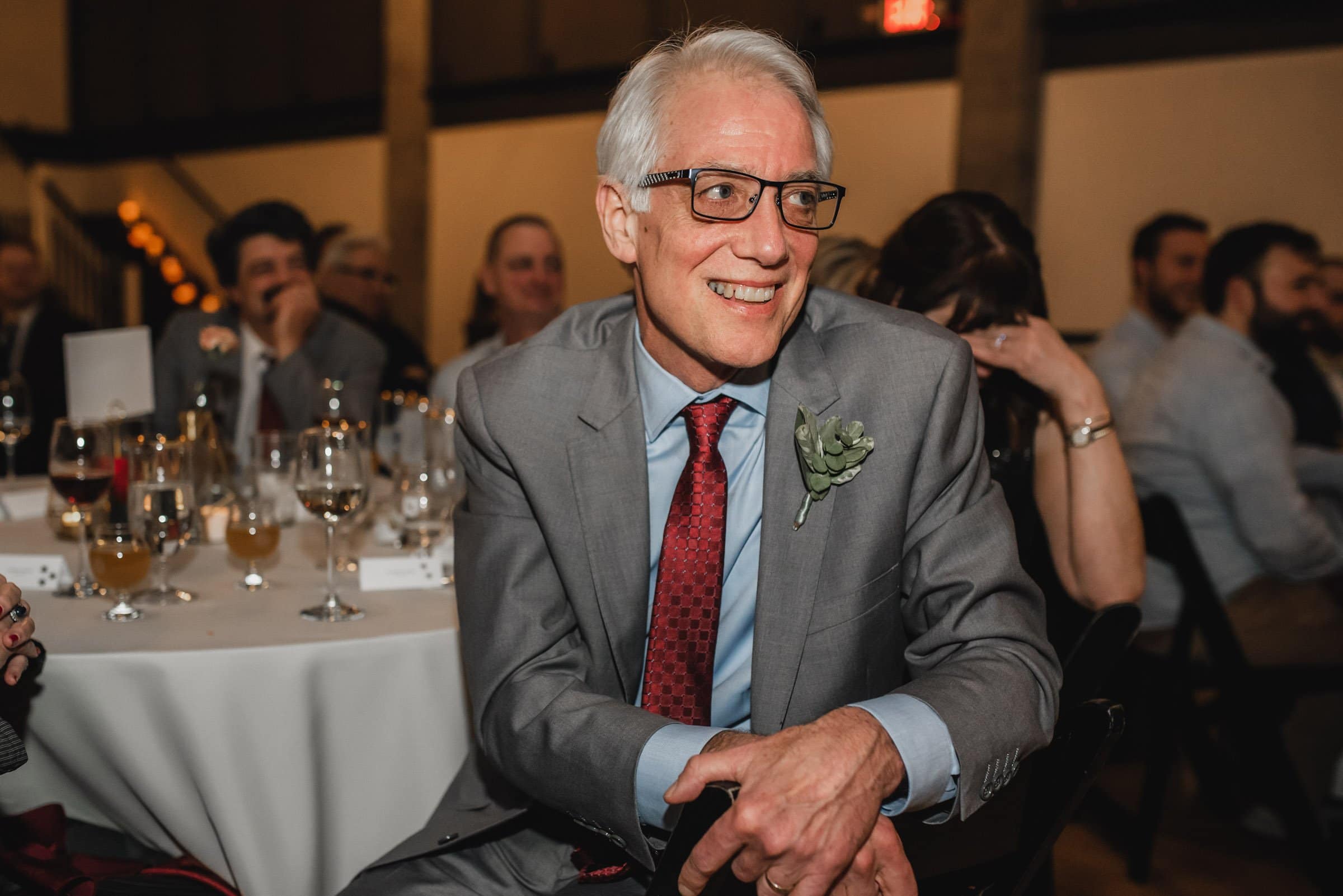 At The Evergreen, a smiling man in a gray suit with a red tie sits at a table during an event, surrounded by people and glasses.