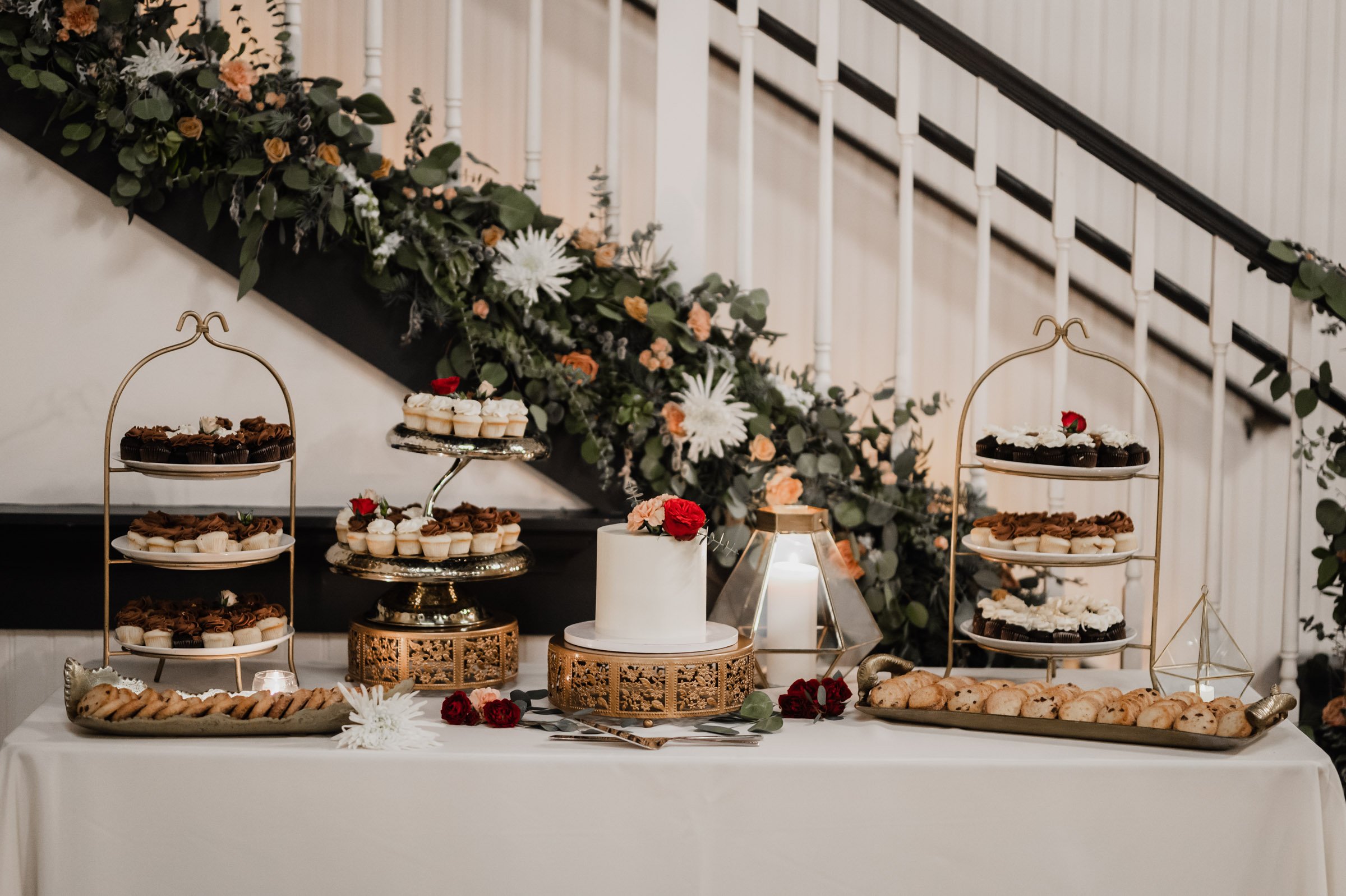 An elegant dessert table showcases a white cake, assorted pastries, and lush greenery in front of a staircase—perfect for capturing wedding photos at The Evergreen.