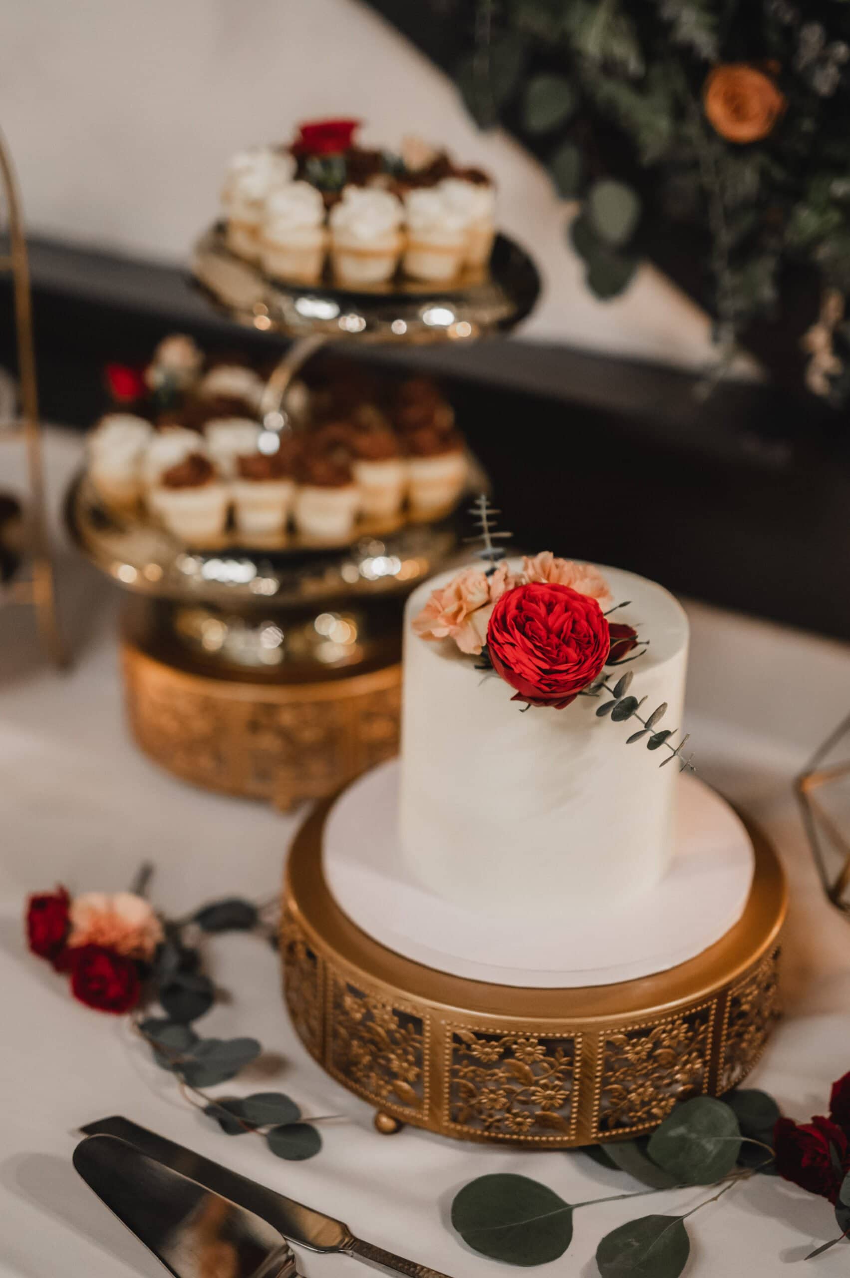 An elegant cake adorned with red and peach flowers sits on a gold stand, surrounded by cupcakes on a tiered tray and scattered leaves. Perfect for capturing stunning wedding photos at The Evergreen.