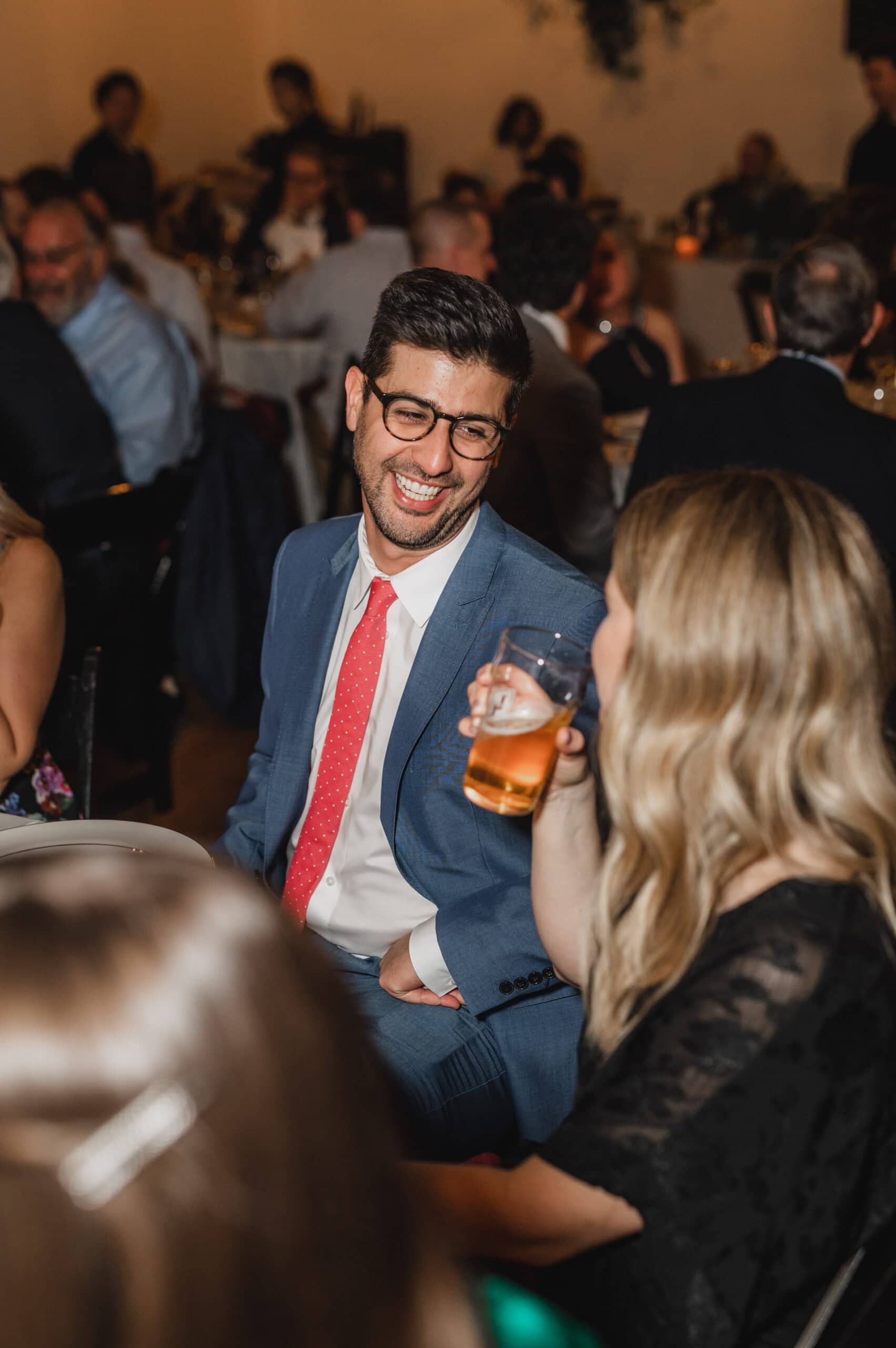 At a crowded indoor event, a man in a blue suit and red tie smiles as he converses with a woman holding a drink. The lively atmosphere is perfect for capturing memorable wedding photos at The Evergreen.