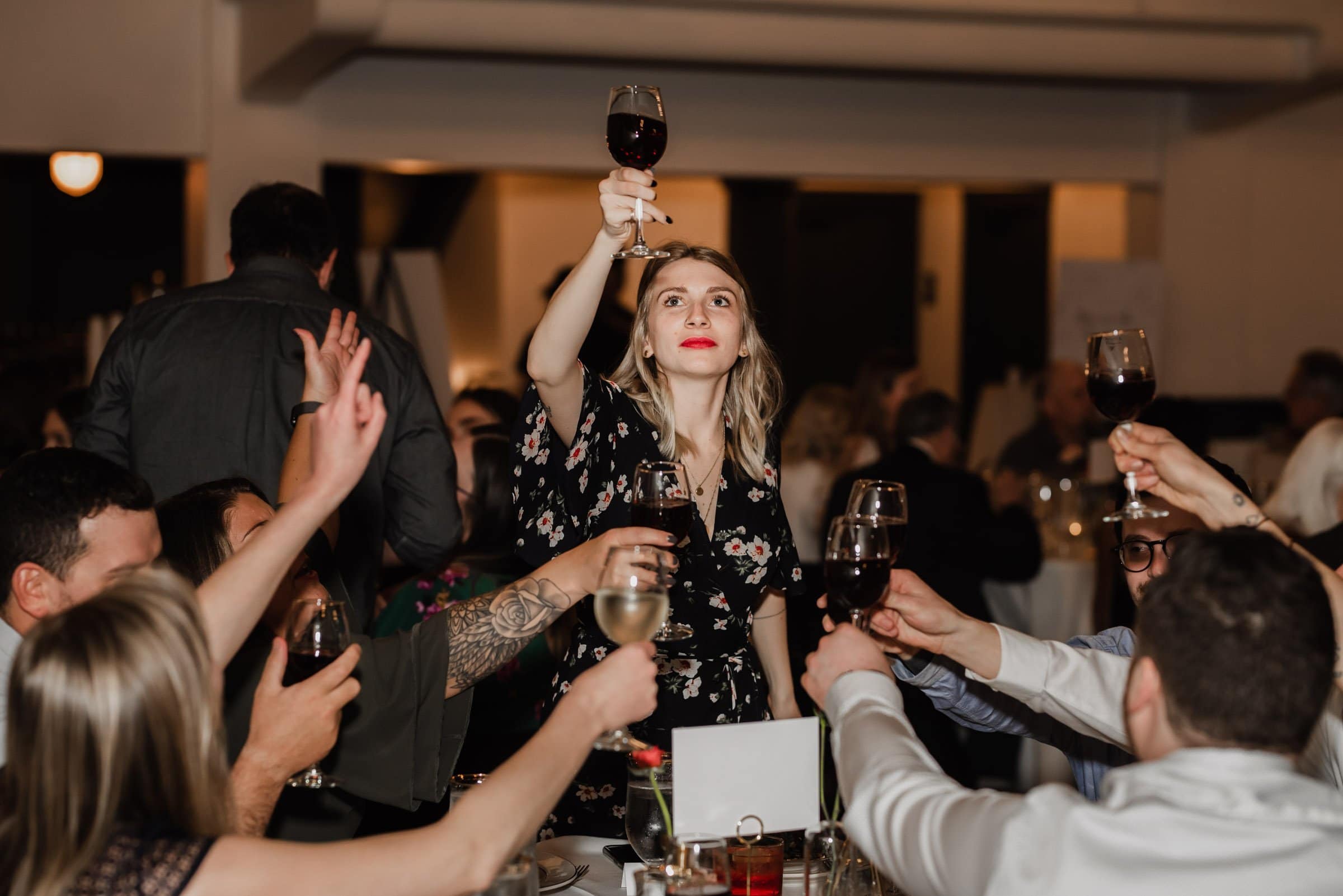 At The Evergreen, a group of people raise their glasses in a celebratory toast during a formal gathering. One woman stands, holding her glass high, capturing the joy often seen in wedding photos.