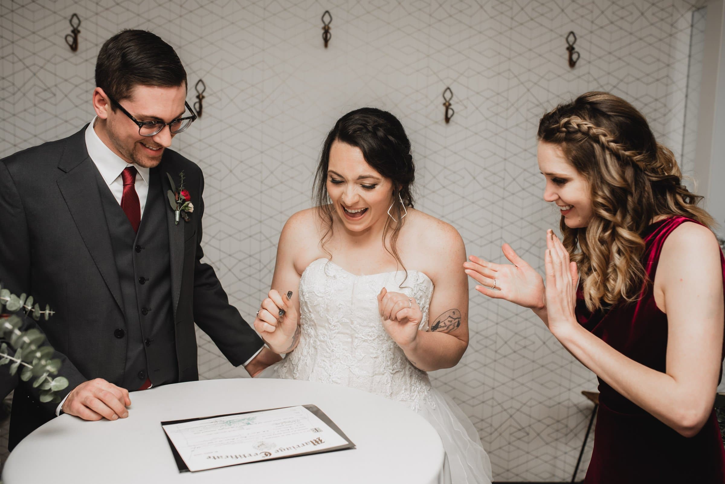 A bride and groom excitedly sign a marriage certificate, with a happy woman clapping beside them as they pose for wedding photos at The Evergreen.