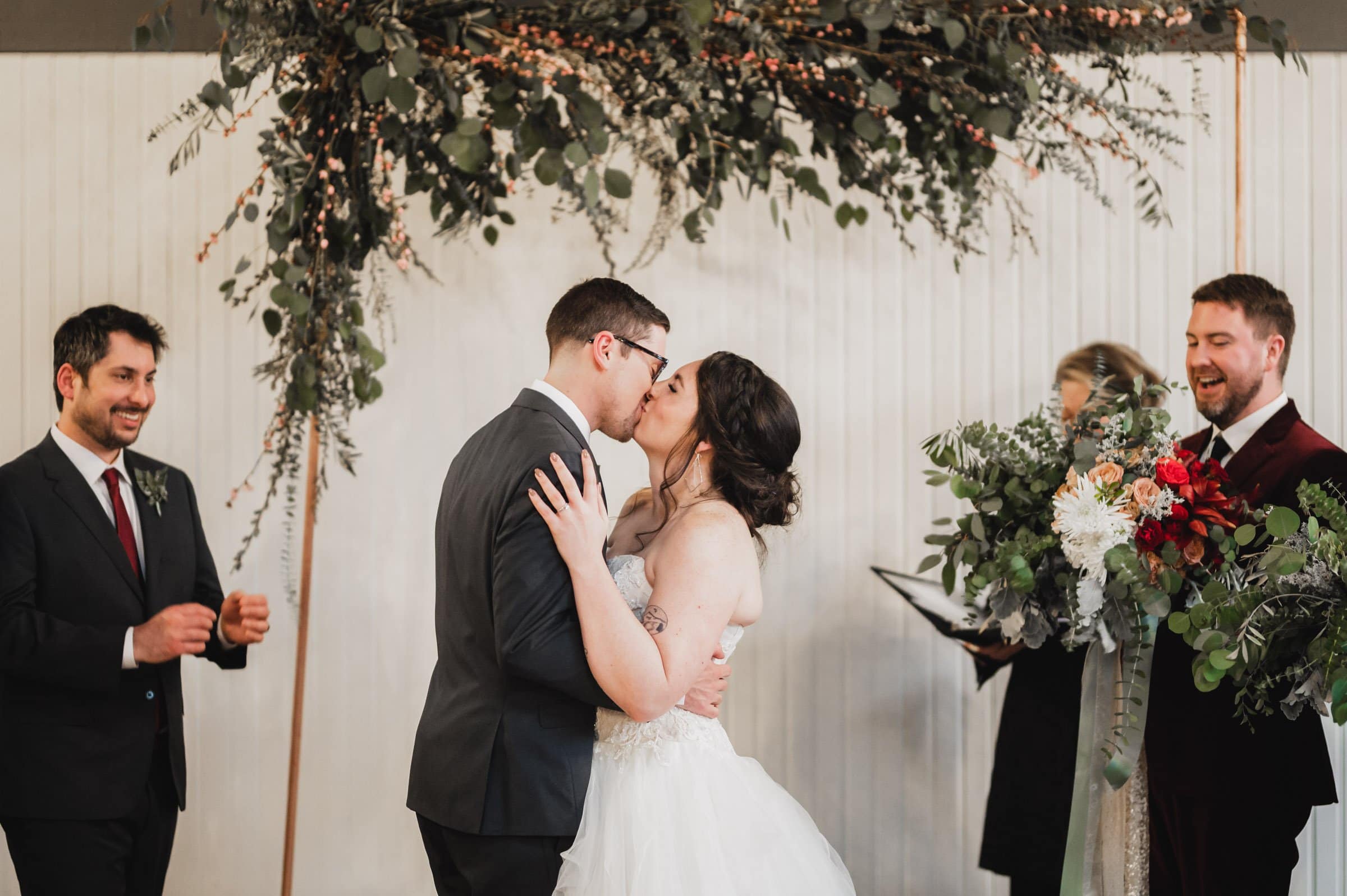 A couple kisses during their wedding ceremony under a floral arch at The Evergreen, surrounded by guests who smile and clap.