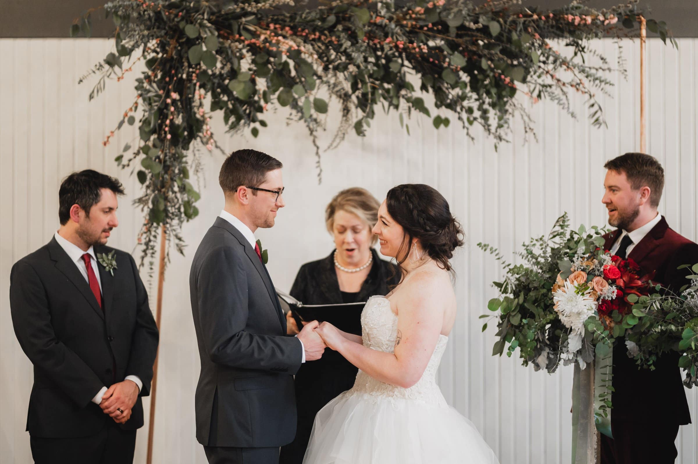 A couple exchanges vows at a wedding ceremony under a green floral arch, with two men and an officiant standing nearby. Capture the moment with stunning wedding photos at The Evergreen.