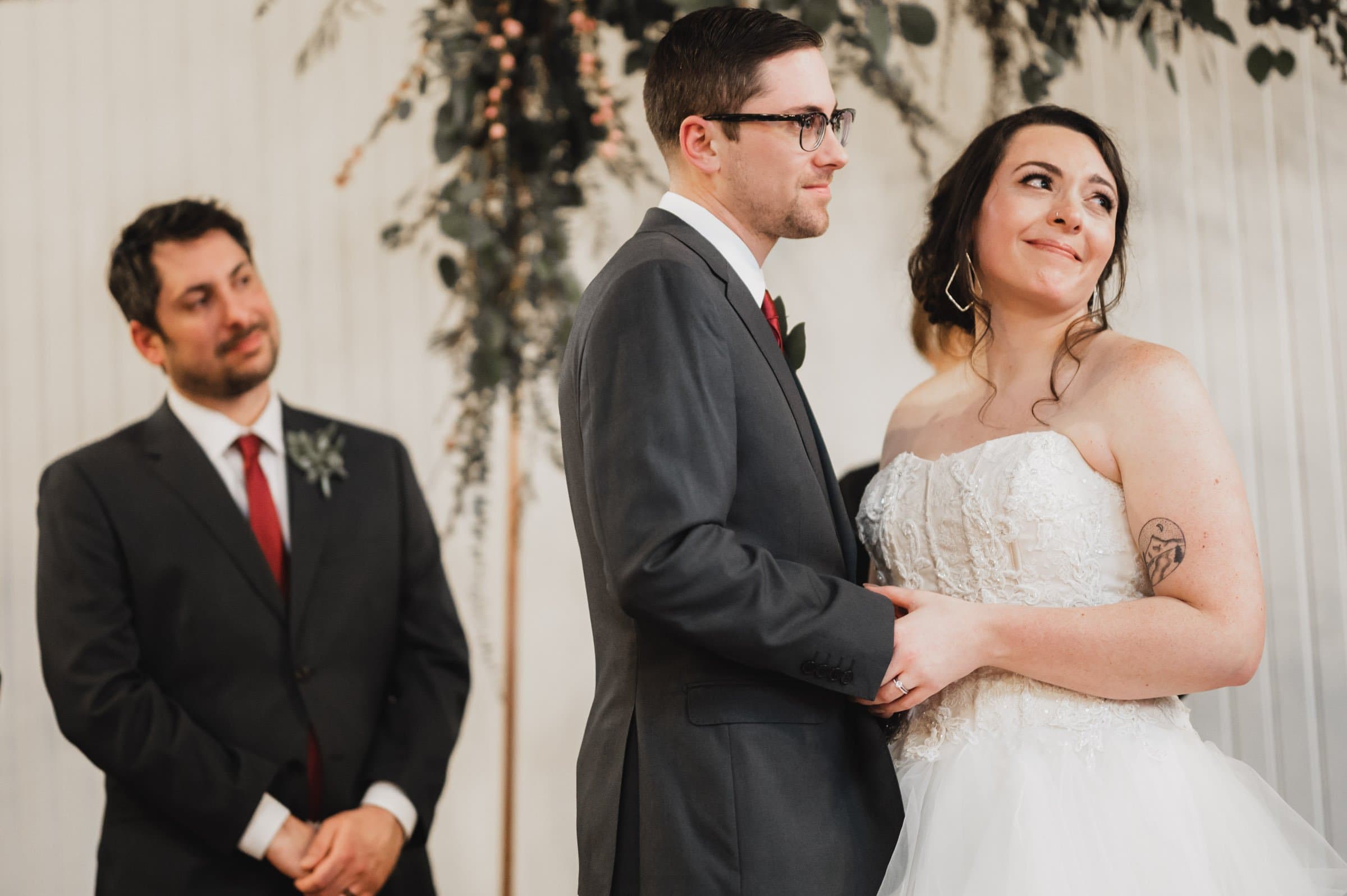 The bride and groom hold hands at their wedding ceremony at The Evergreen, with a groomsman watching them closely, capturing a perfect moment for their wedding photos.