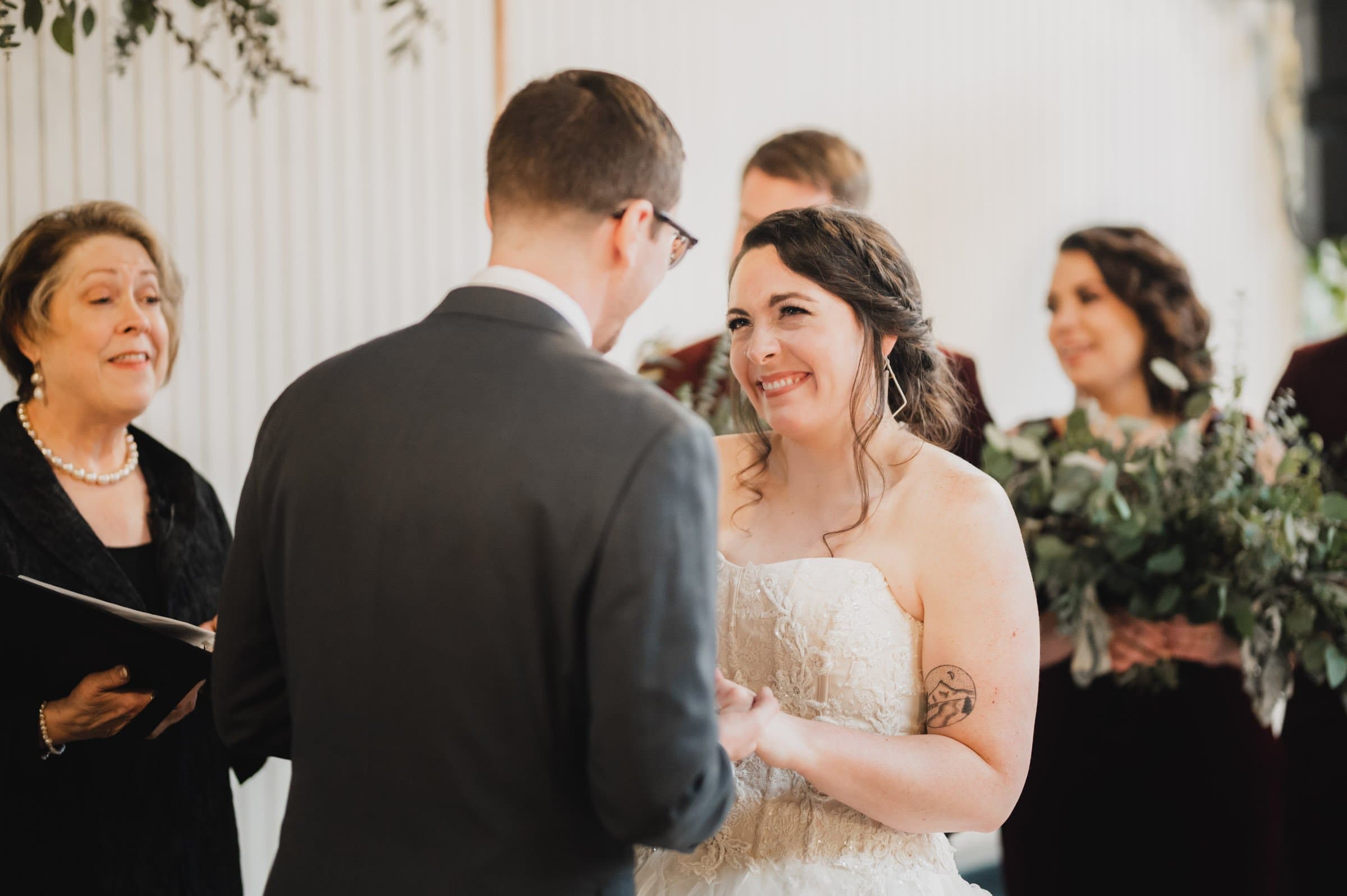 The bride smiles at the groom during their wedding ceremony, surrounded by bridesmaids and the officiant. Captured among the stunning backdrop of The Evergreen, these cherished moments will be treasured forever.