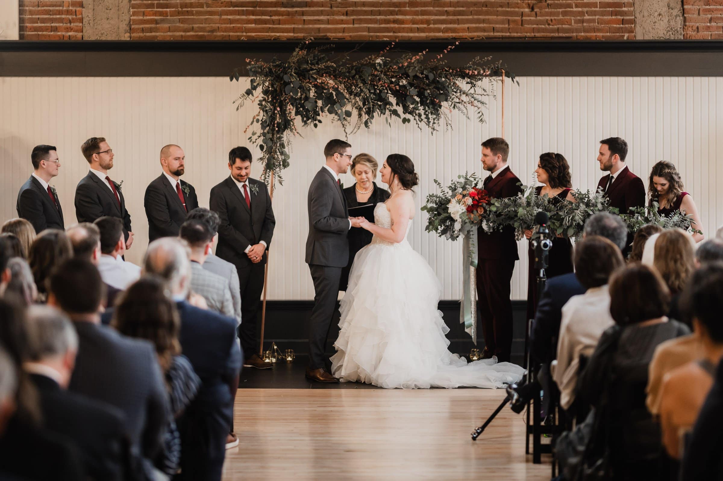 At The Evergreen, a bride and groom exchange vows during an intimate indoor wedding ceremony, surrounded by their bridal party and cherished guests.
